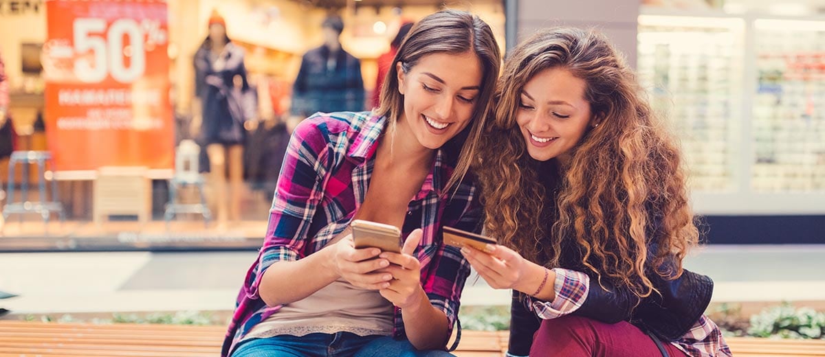 Two young women sitting on a bench, one looking at her phone and the other holding a credit card.